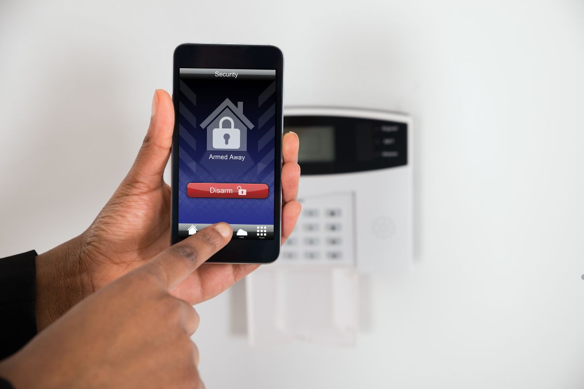 A person using a smartphone app to disarm a home security system, with a keypad visible on the wall behind.