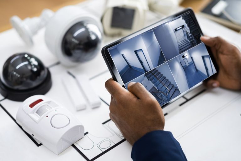 A person holds a tablet displaying security camera feeds, surrounded by surveillance equipment and architectural plans.